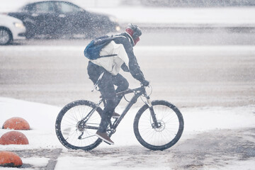 Man riding bike on snowy city street during snow storm. Cyclist rides on snowy road during blizzard. Man in warm clothes riding on slippery road on bicycle. Cyclist while driving on snow-covered road