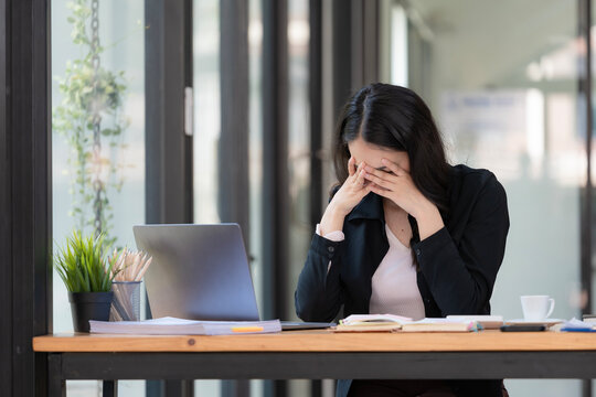 A tired businesswoman under stress works at a laptop while sitting at a table at home and holds her hand on her temples, causing migraine attacks.