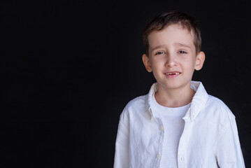 Portrait of a 6-year-old boy in a white shirt against a black wall. Smiles widely, one front tooth is missing, fell out. 