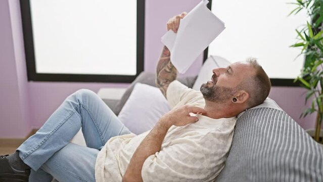 Hot weather torment, young man fanning himself with documents, suffering in a windless room, sitting on sofa at home
