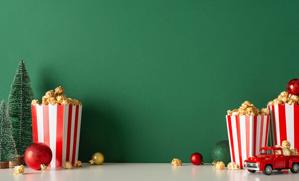 Enjoy cozy Noel celebration at home with popcorn delivery theme. Side-view image of table featuring tiny red car, popcorn in boxes, baubles, small fir against green wall backdrop, room for movie promo