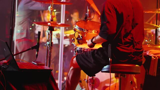 Back view of a man playing drums. A drummer is performing on stage during a concert. An orchestra and a band conductor are in the background. Close-up. A drummer surrounded by light effects