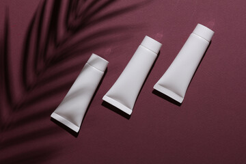 White cream tubes awith on burgundy background with palm leaf shadow. Creative mockup