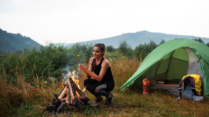 Woman squatting near green tent and warming hands over bonfire