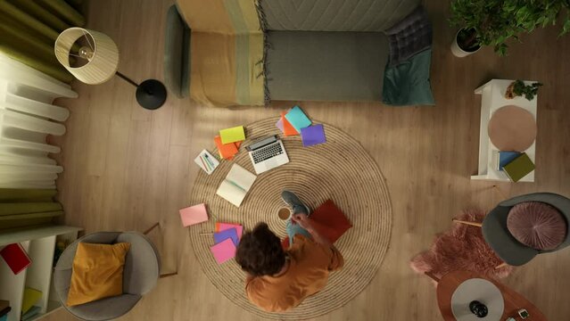 Top view capturing a young man sitting on the floor and studying, working on a laptop and taking notes, surrounded with colorful books.