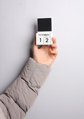 Man's hand in warm down jacket holds block calendar with date november 12 on gray background