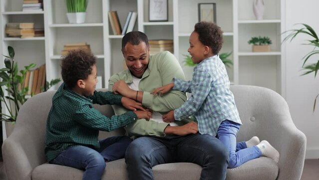 Cheerful little boys tickling on sofa while playing with father at home. Happy black family enjoying weekend at home.