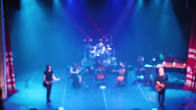 Rock band and small symphony orchestra playing world rock hits. Front view. Soft focus. Colourful light show accompaniment. With band, violin, cello, piano, guitar, bass, drums