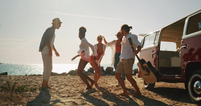 Slow Motion Portrait of Group of Young Friends Sitting Next to a Minivan and Celebrating by Dancing and Singing on a Beach. Multiethnic Men and Women Having Fun, Enjoying Their Youth and Summer Days