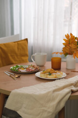 Autumn Home breakfast, vaze with yellow leaves,candle, grey plate with fried egg, sausage, bean, cup of coffee, cottage cheese with peach on round wooden table..