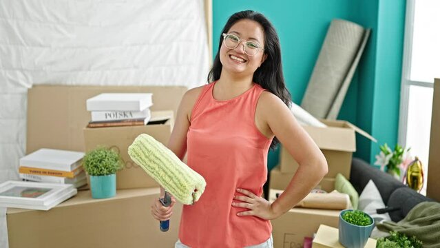 Young chinese woman smiling confident holding paint roller at new home