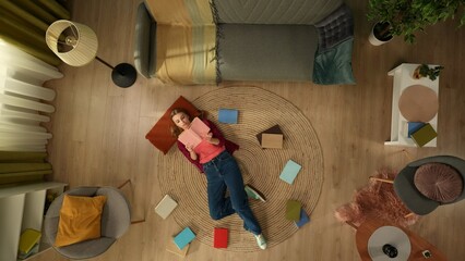 Top view capturing a young woman laying on the floor of her room, flipping pages of the book, reading patiently.