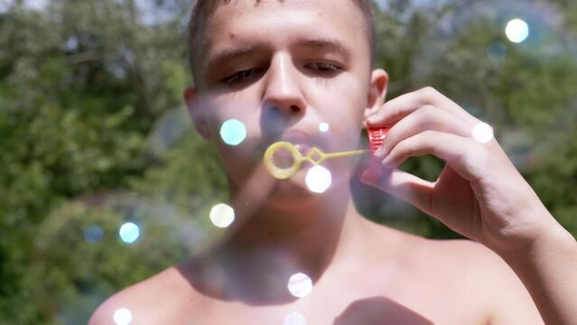Boy with a Naked Torso Blowing Soap Bubbles in Nature in the Rays of Sunlight. Close up. A teenager is relaxing outdoors in the forest, blowing colorful bright soap bubbles. Blurred green background.