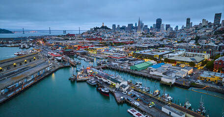 Fishermans Wharf at dusk with night lights on over city and pier with Oakland Bay Bridge aerial