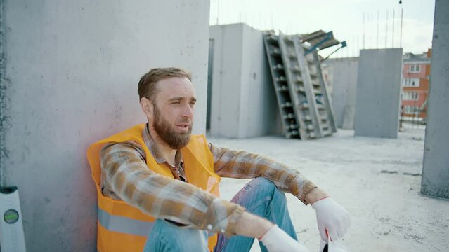 Young tired man worker sitting and takes off white hardhat at construction workplace. Slow motion
