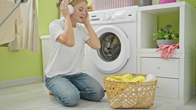 Young blonde woman listening to music holding clothes of basket at laundry room
