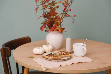 Home autumn breakfast, two toasts with almond butter, figs, coffee, pumpkins, candle, ceramic vase with red leaves on a round wooden table