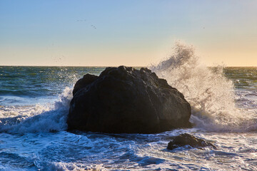 Sunlight behind wave crashing behind dark boulder surrounded by sea foam near sunset © Nicholas J. Klein