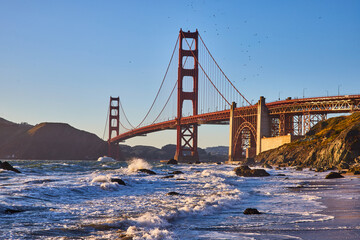 Waves rolling into shore of sandy beach with view of Golden Gate Bridge at sunset