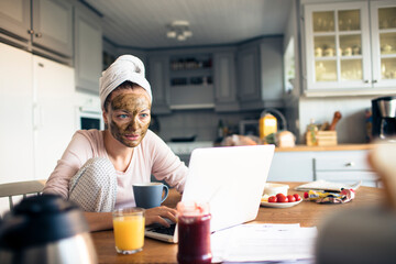 Attractive young woman with a facial mask using a laptop in the kitchen