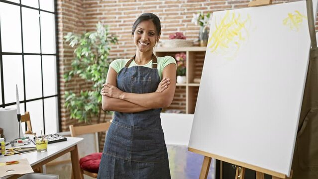 Young latin woman artist standing with arms crossed gesture smiling at art studio