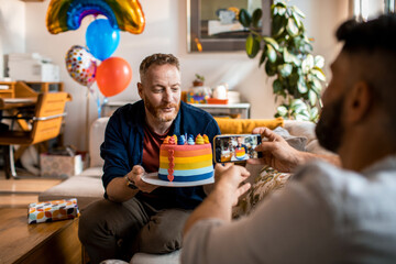 Young gay man holding a birthday cake and getting his photo taken by his partner at home