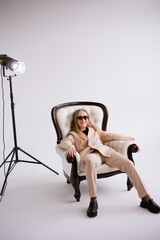 Female, model, blogger, blonde in sunglasses sitting on chair in white photo studio, isolate. Dressed in a formal beige trouser suit