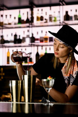 Young attractive woman bartender carefully pours drink from crystal bottle into steel jigger