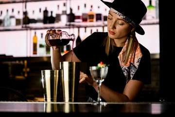 Young woman bartender pours red alcoholic drink into shaker to make cocktail at bar counter