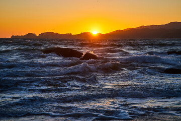 Choppy waves with boulders in water and golden sky with sun poking above mountains at sunset