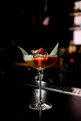 Martini glass of tasty cocktail decorated with banana leaf, ice cube, raspberry and citrus zest plane standing on barstand