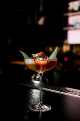 Martini glass with brown alcoholic drink decorated with green leaf, berry and citrus zest stands on bar counter