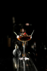 Glass with brown alcoholic drink decorated with green leaf, berry and citrus zest stands on bar counter