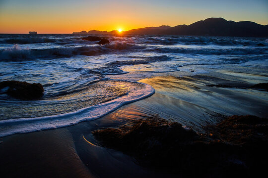 Sunset over distant mountains with blue waves crashing into sandy beach