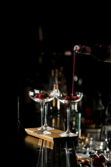 Barman's hand holds bottle with red syrup and pours it into three martini glasses decorated with berries at bar counter