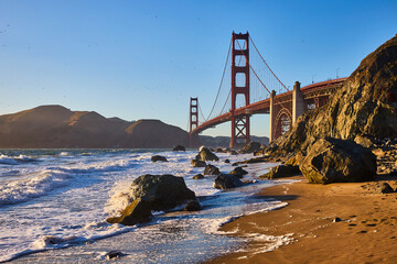 Waves rolling into sandy beach with massive boulders and Golden Gate Bridge near sunset