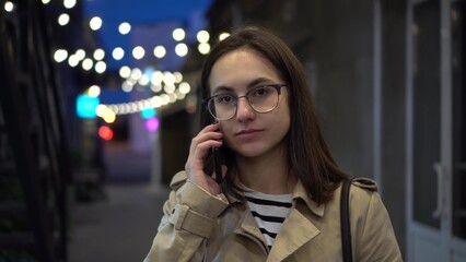 A young woman talks on the phone late at night on a narrow street. A girl with glasses communicates on a mobile phone.