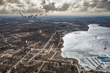 Overhead view of Victoria Harbour, Ontario, during winter. Geese fly above frozen landscapes while windsurfers glide on icy waters. Cloudy skies shadow the quaint town with snow-kissed roofs.