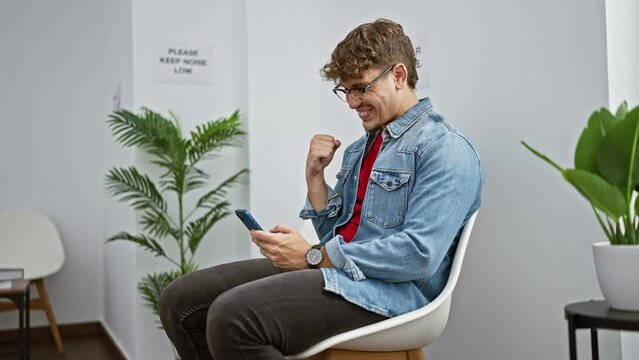 Jubilant young hispanic man sitting in a waiting room chair, texting a victorious message on his smartphone amongst indoor background