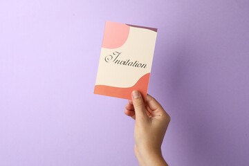 Woman holding beautiful card with word Invitation on lilac background, closeup