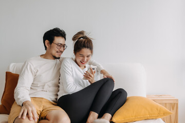 Happy lovely asian couple sharing smartphone to watch together in living room.