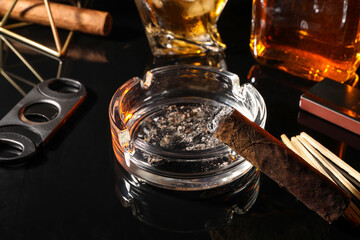 Burnt cigar, ashtray, cutter and whiskey on black mirror surface, closeup
