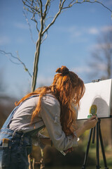 A beautiful female artist is painting a picture on a easel while standing at the park in nature