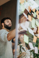 Diverse team brainstorming, discussing goals, and collaborating in a modern office. Using sticky notes on a glass wall for planning and strategizing, they showcase teamwork and persistence.