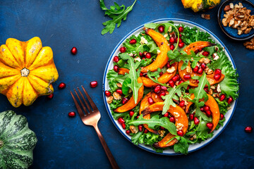Vegan autumn salad with baked sweet pumpkin, lettuce, arugula, pomegranate seeds and walnuts. Comfort slow  food. Blue background. Top view