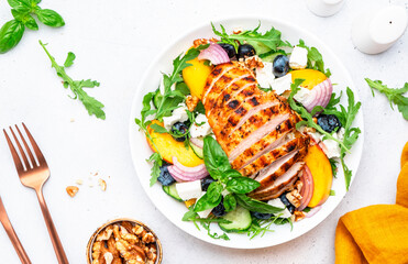 Tasty salad with peaches, grilled chicken breast, blueberries, red onion, feta cheese, basil and arugula, white table background, top view