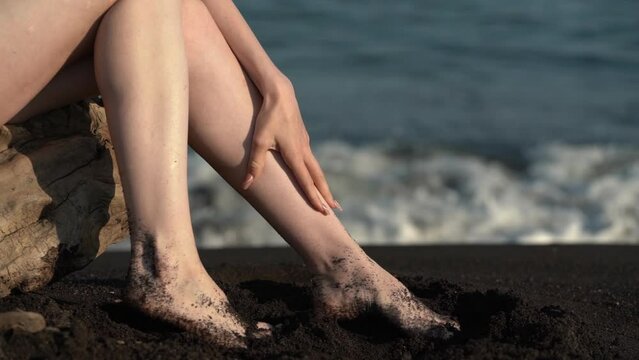 Close-up of bare female feet on sand on beach. Woman stroking legs on background of sea waves in sunny day. Part of female body with long legs sitting on black color sandy beach and touching herself