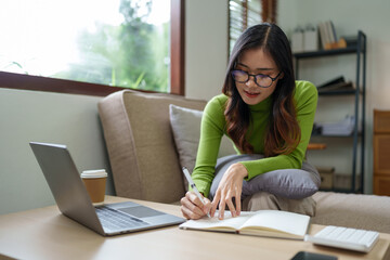 Young successful professional Asian businesswoman in casual clothes sits on a sofa, reads a book, writes notes in a notebook while working on a laptop computer at home office in the living room.