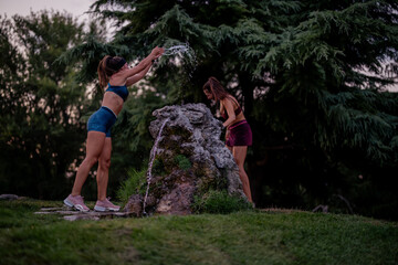 Fit, athletic women enjoying evening workout in park. Having fun playing with water, splashing each other and laughing. Active and healthy sporty lifestyle captured in photo.