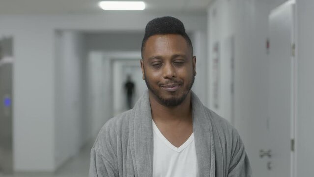 Happy African American man stands in hospital corridor, smiles and looks at camera. Adult patient during treatment in modern clinic. Medical personnel walks in the background. Slow motion. Portrait.
